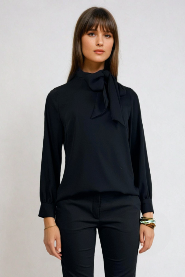 A woman wearing a black tunic blouse with long sleeves and a bow tie at the neck, paired with black slim-fit pants. Accessories include black shoes and a watch.